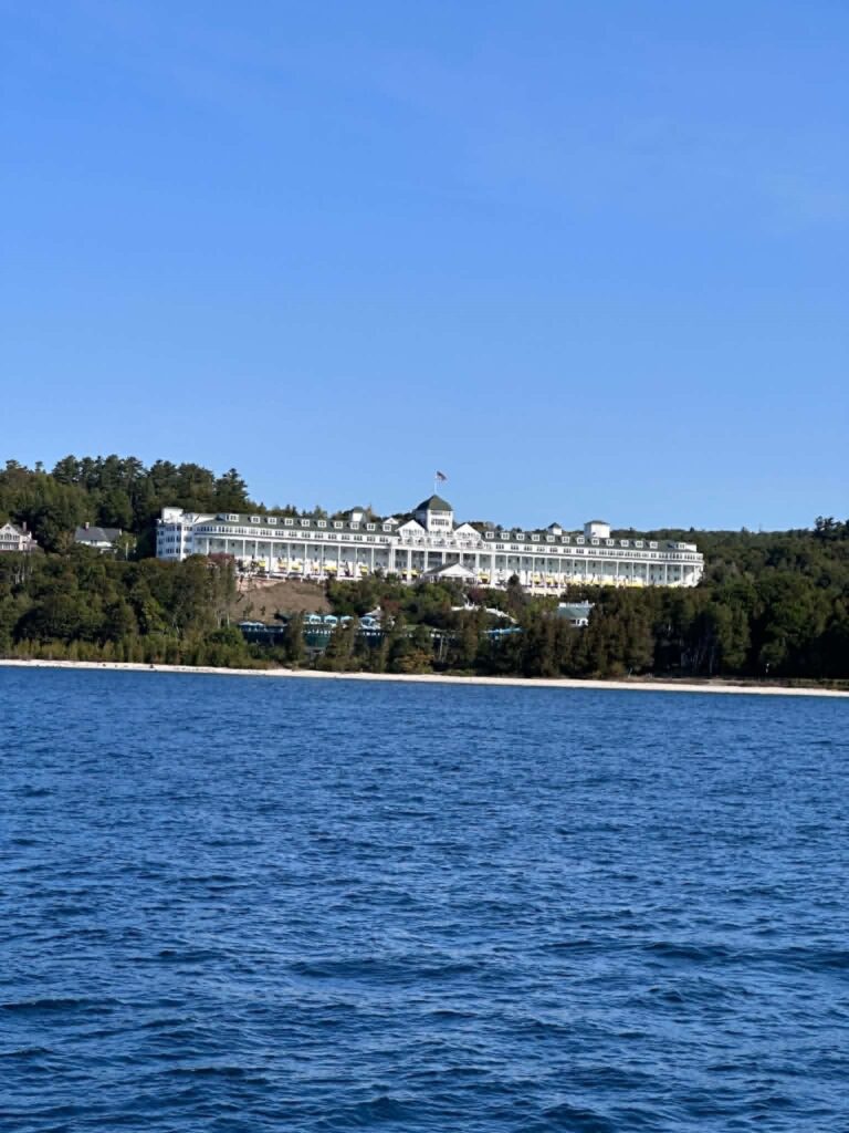 The full white facade and long columned porch of the Grand Hotel on Mackinac Island, with its green-domed cupola and flag, seen from Lake Huron