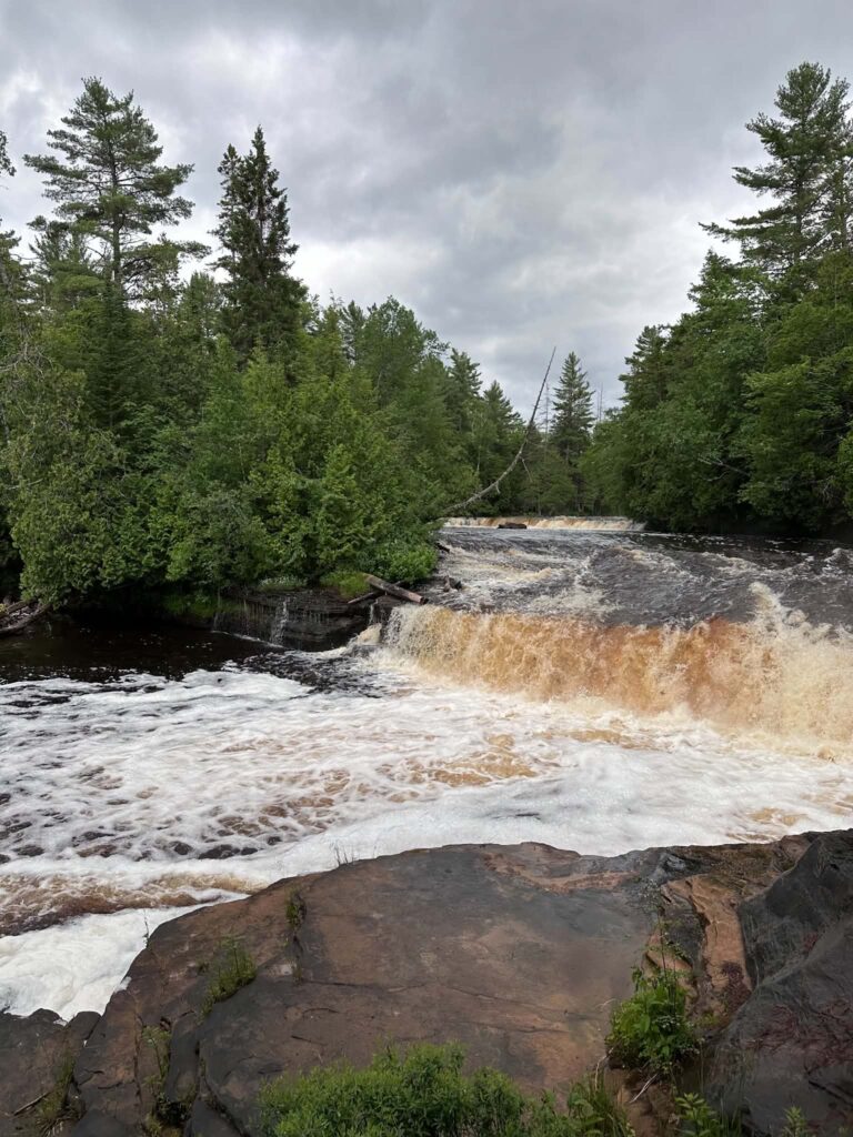 Amber-colored water rushing over one of the Lower Tahquamenon Falls cascades with white foam at the base
