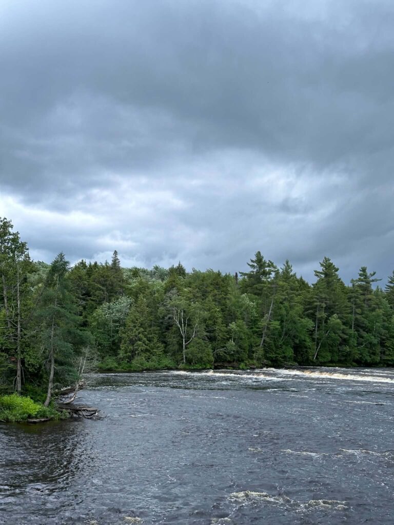 Calm wide bend of the Tahquamenon River approaching the Lower Falls under a dramatic overcast sky with lush green shoreline