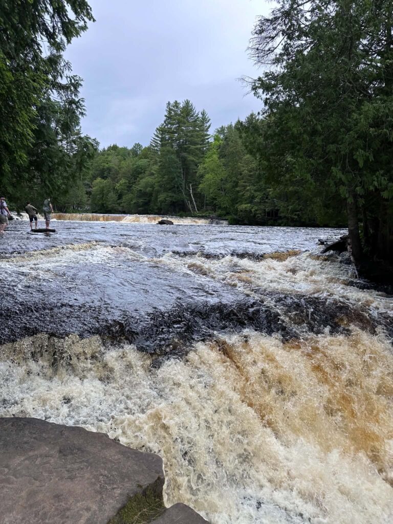 Amber water rushing over a curved rock ledge at the Lower Tahquamenon Falls with white foam churning at the base