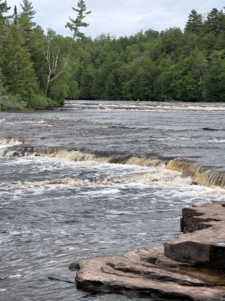 Flat exposed rock shelf at water level where the Tahquamenon River spreads wide across the Lower Falls area