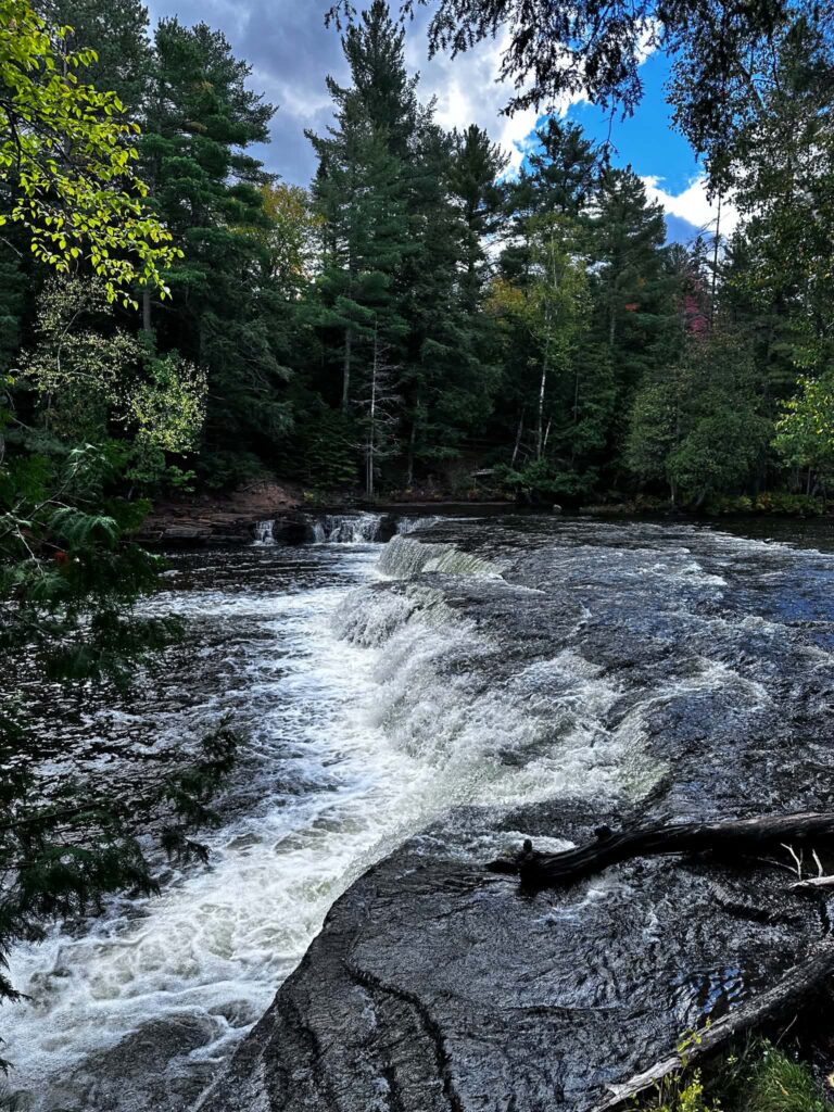 Lower Tahquamenon Falls at low water with a shallow cascade spreading gently over flat rocks with green forest in the background