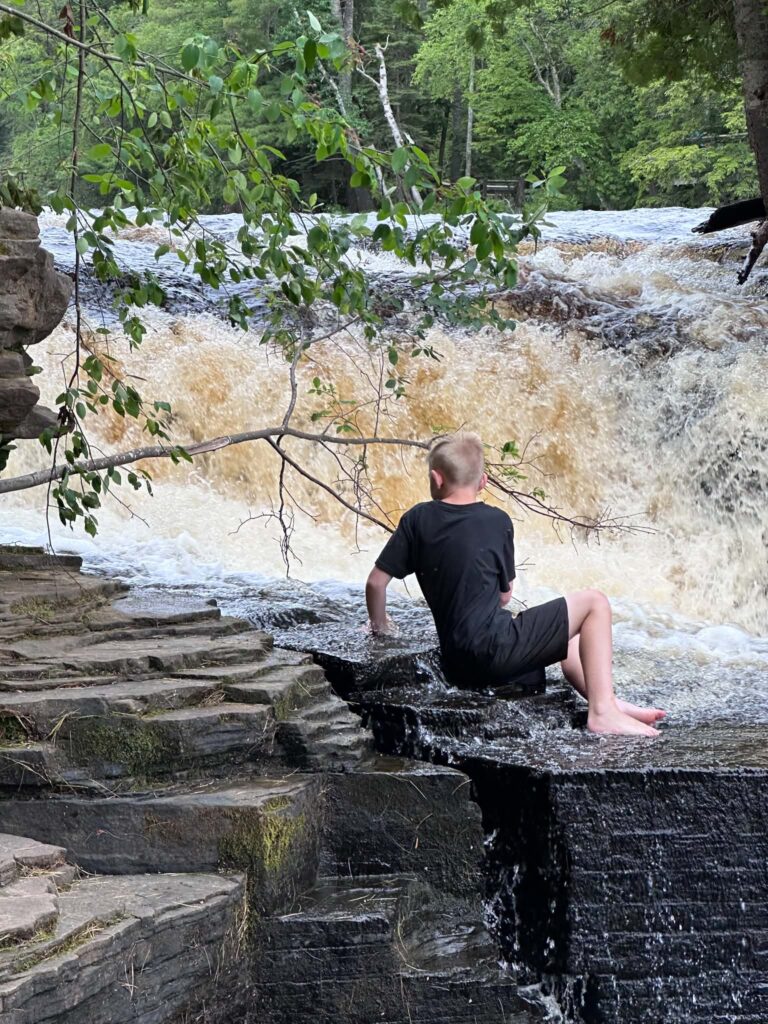A visitor sitting on a flat rock at the edge of the Lower Tahquamenon Falls with a rushing cascade just beside them