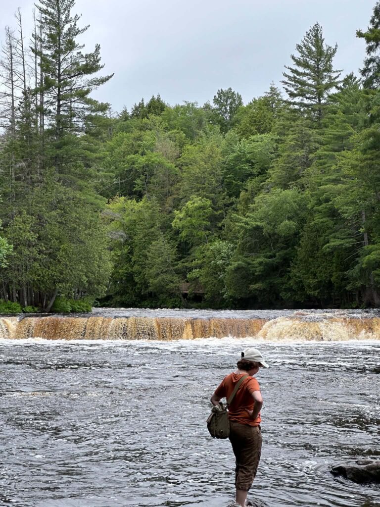 A visitor standing on a rock beside a Lower Tahquamenon Falls cascade, giving a sense of the waterfall's scale against the surrounding forest