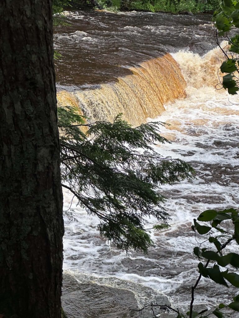 Portrait-view of an amber-colored Lower Tahquamenon Falls cascade dropping between dark rock walls in dramatic lighting