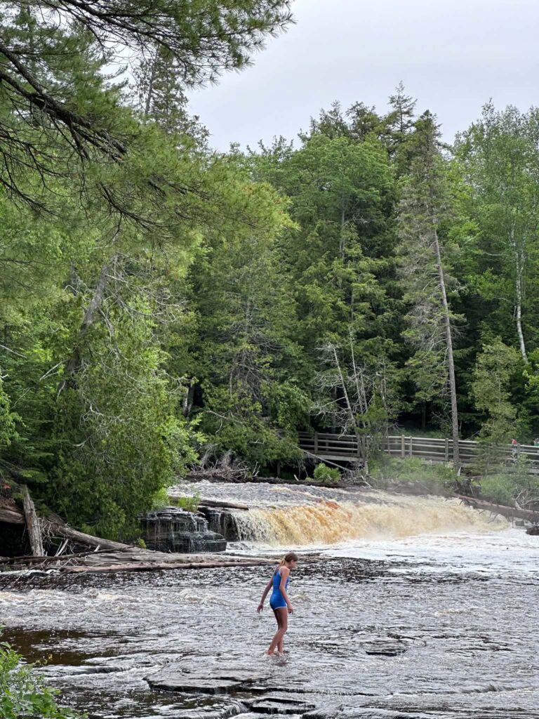 A visitor wading in the shallow amber water at the Lower Tahquamenon Falls on a summer day with cascades visible in the background