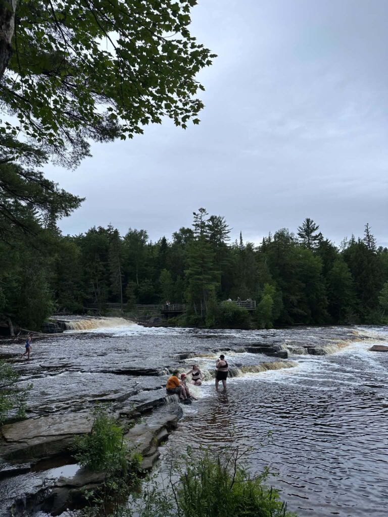 Visitors standing and sitting on the flat sandstone rocks beside a rushing cascade at the Lower Tahquamenon Falls