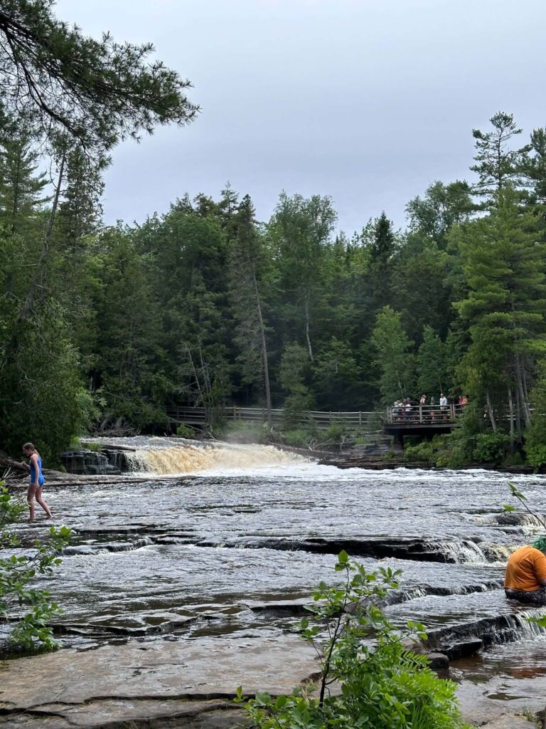 Visitors resting on rocky ledges at the Lower Tahquamenon Falls with an amber-colored cascade rushing behind them