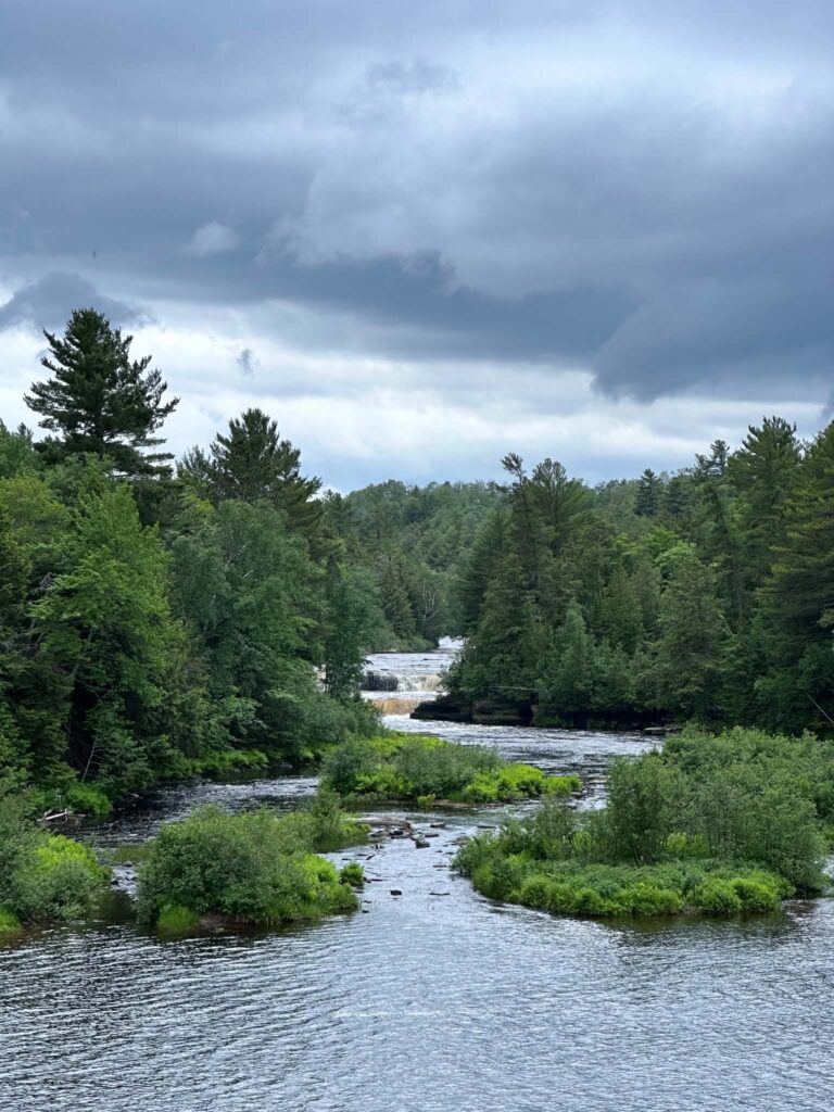 Wide calm stretch of the amber-colored Tahquamenon River approaching the Lower Falls under cloudy skies