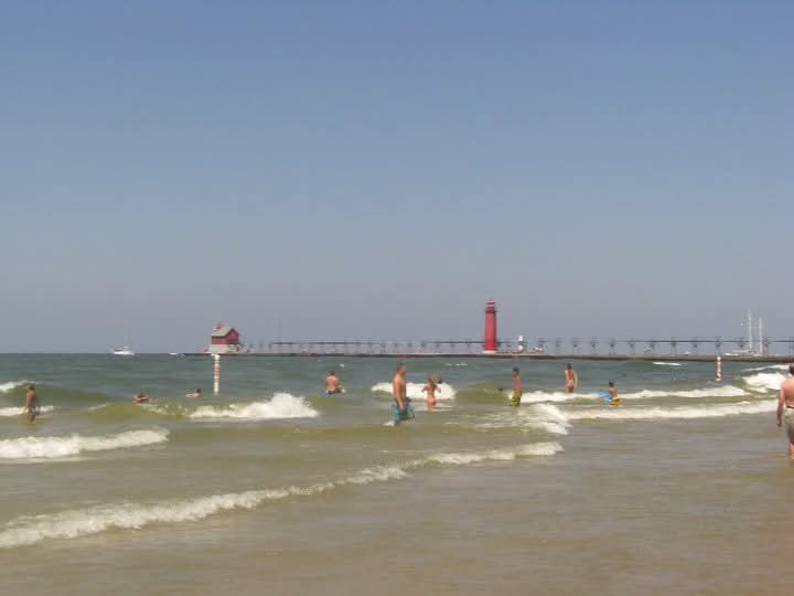 Grand Haven State Park beach — swimmers in the waves with the iconic pier and red lighthouse beyond