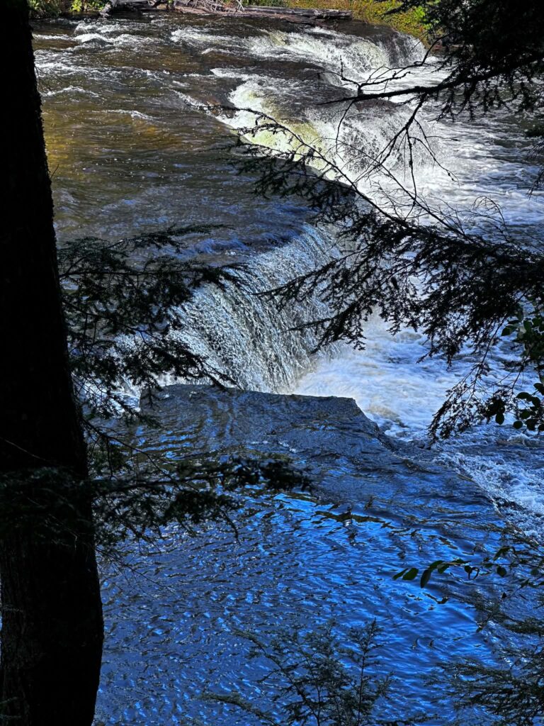 The Lower Tahquamenon Falls cascade over flat rocks on a sunny day with vivid blue sky overhead and green summer forest behind