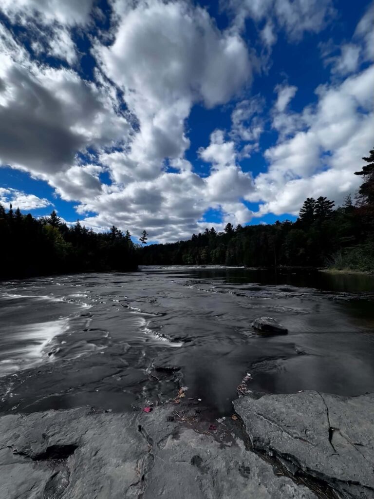 Dramatic wide-angle view of the Lower Tahquamenon Falls area with a vast blue sky and billowing clouds above the exposed flat sandstone rock shelf