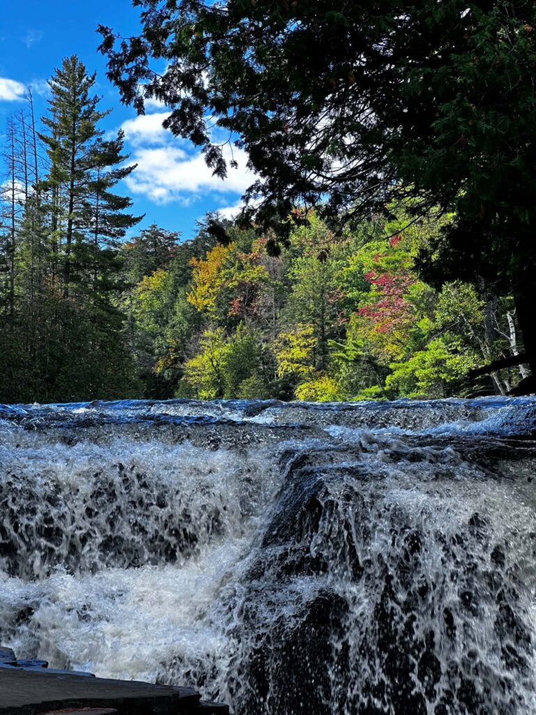 Lower Tahquamenon Falls on a sunny autumn day with brilliant fall foliage in orange, gold, and green above the flat rock shelf and blue sky overhead