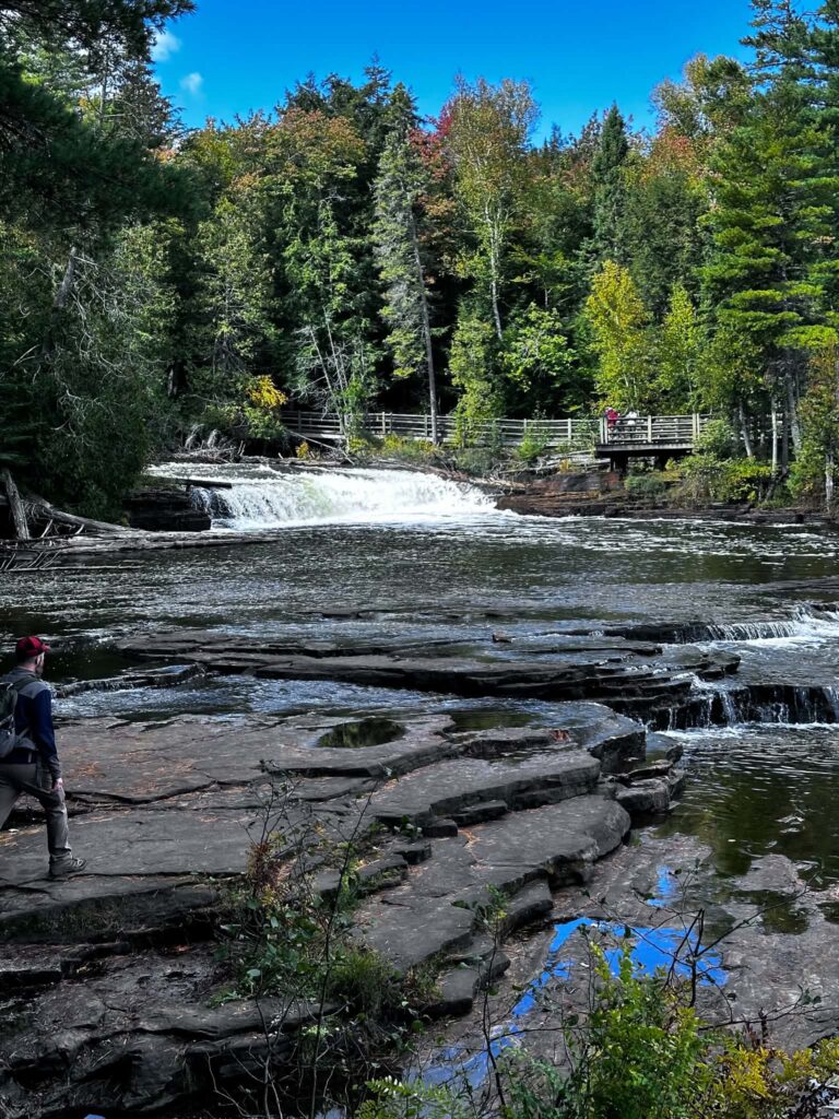 Wide flat sandstone rock shelf at the Lower Tahquamenon Falls with a thin cascade at the far edge and fall-colored trees in the background
