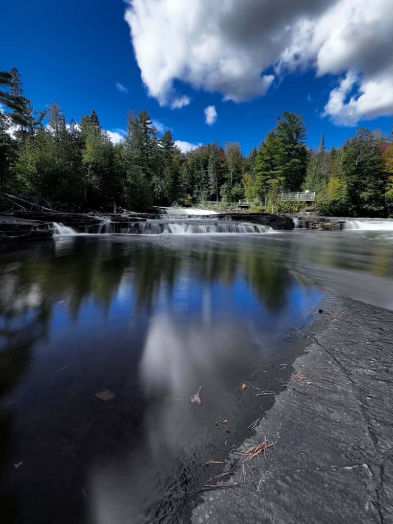 Sunny summer view of the Lower Tahquamenon Falls area with a gentle cascade, calm river, and vivid blue sky with scattered clouds