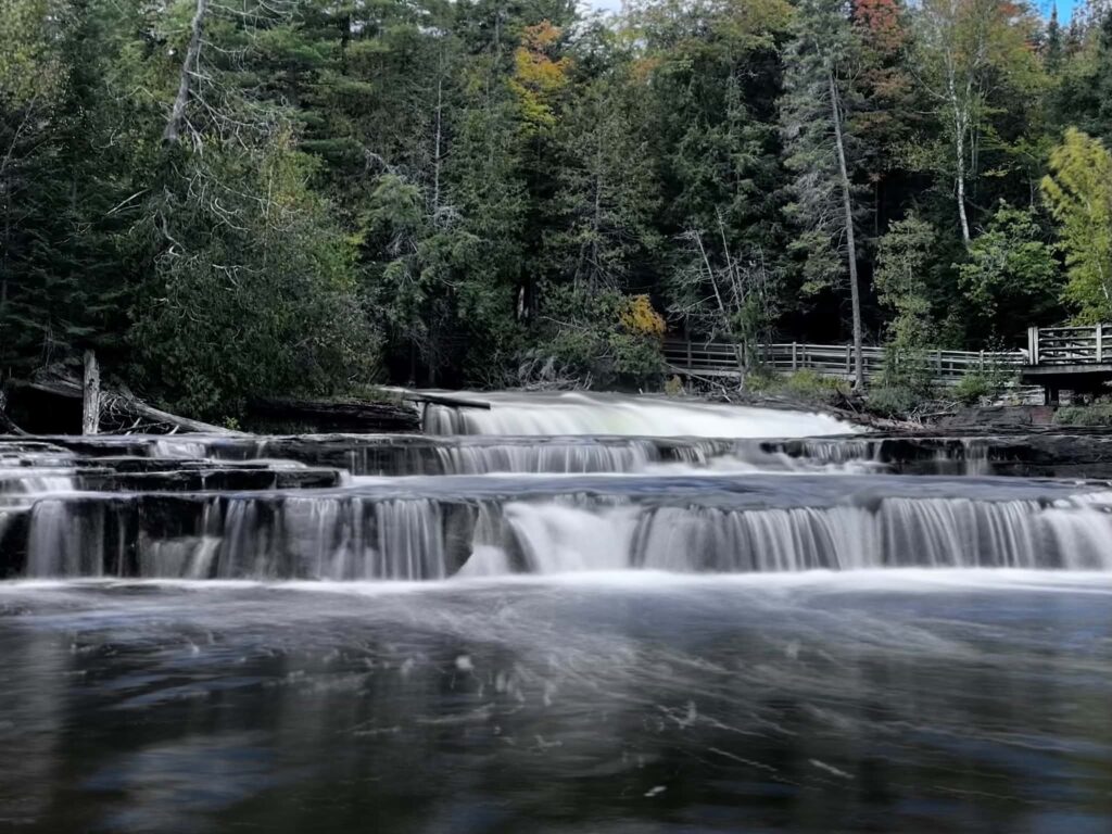 Wide multi-step cascade of the Lower Tahquamenon Falls flowing over flat sandstone rocks under an overcast sky