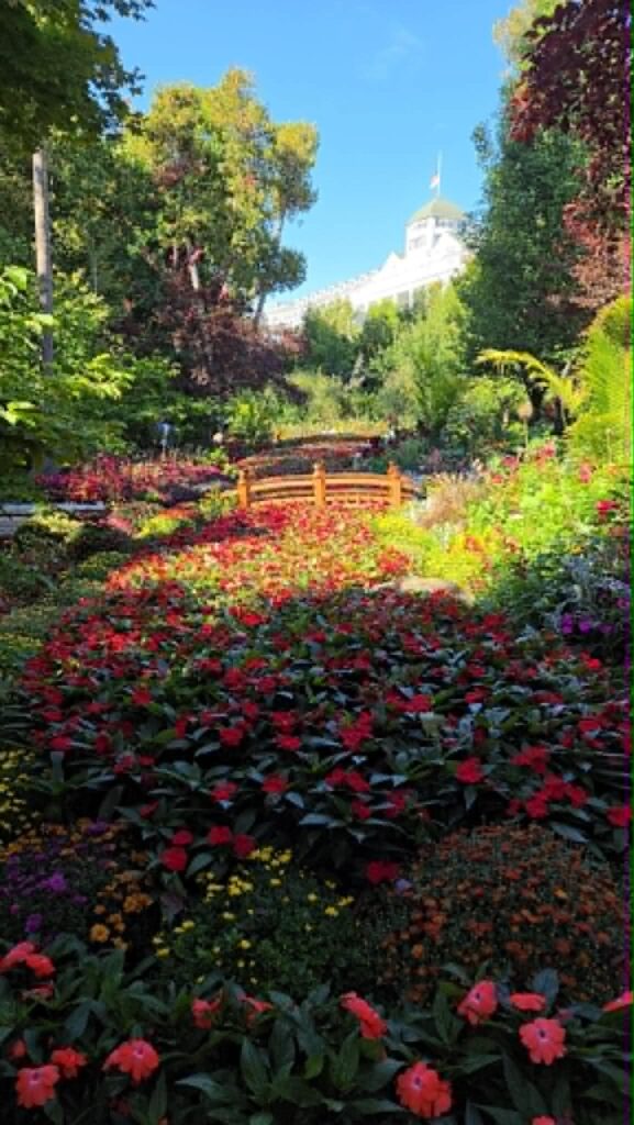 Grand Hotel partially visible beyond a colorful flower garden and wooden footbridge on Mackinac Island.
